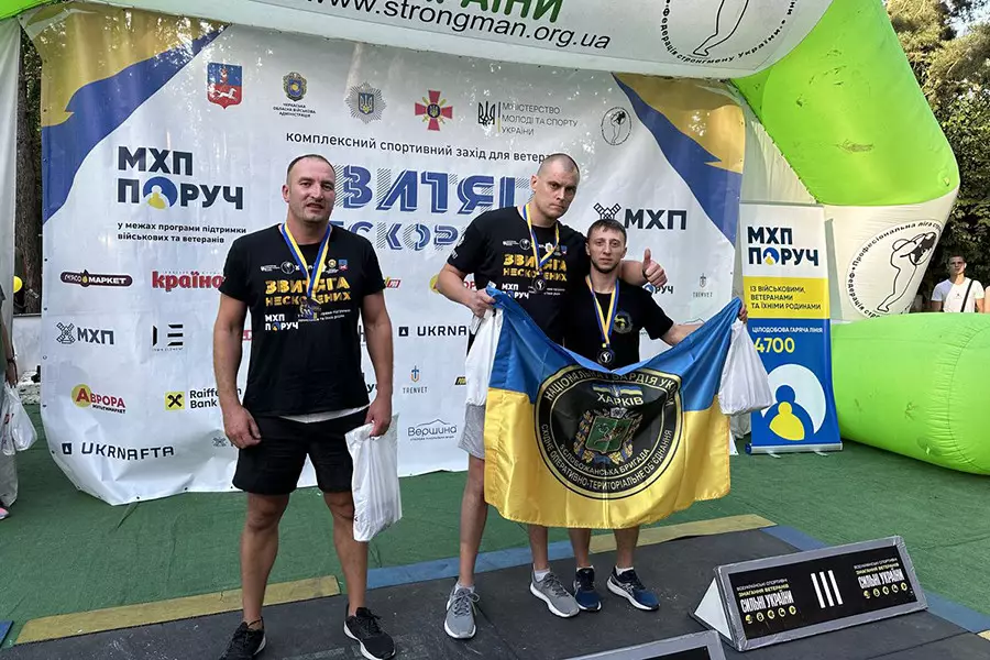 Три чоловіки-переможці із медалями на фоні банеру турніру, двоє з них тримають прапор Бригади